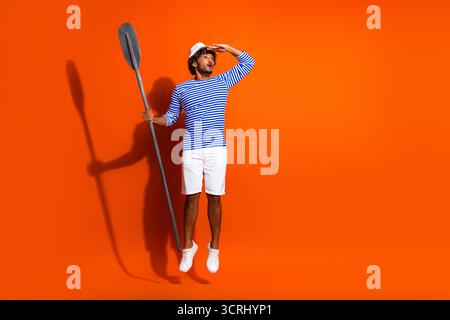 Junger Mann in einem von Seemännern inspirierten gestreiften Hemd mit Paddel auf leuchtendem orangefarbenem Hintergrund, das Sommerreisen und Abenteuer zeigt Stockfoto