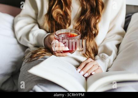 Frau, die eine Tasse Tee hält, während sie Buch liest. Gemütliche Lifestyle-Szene zu Hause. Warmes Getränk und weiche Decke in ruhiger, entspannter Atmosphäre. Stockfoto