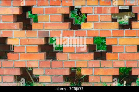 Eine rote Ziegelwand mit gleichmäßig verteilten Lücken zeigt grüne Grasflächen und Pflanzen dahinter und schafft einen geometrischen Kontrast zwischen Architektur und nat Stockfoto
