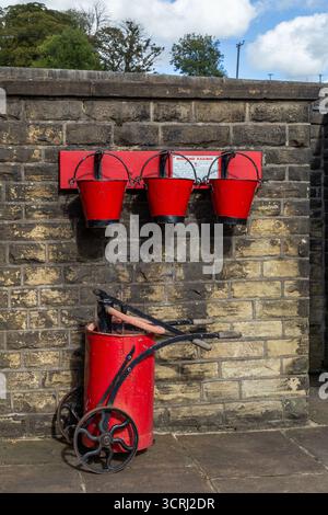Historische rote Feuereimer und ein roter Wagen voller Ausrüstung an der Oakworth Station, Teil der Keighley and Worth Valley Railway in Yorkshire. Stockfoto