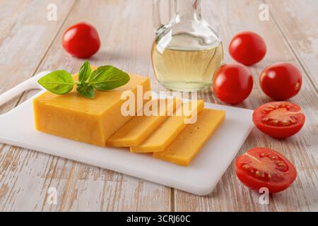Cheddar-Käse und Tomaten aus nächster Nähe. Ganzer Käse auf weißem Schneidebrett und rote Tomaten auf rustikaler Holzplatte. Vorderansicht Stockfoto