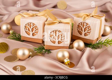 Beige und braune Geschenkboxen mit goldenen Bändern auf beigefarbenem Seidenhintergrund. Geschenkboxen, Weihnachtskugeln, Tannenzweige und rundes Konfetti. Stockfoto