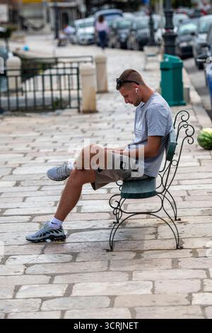 Junger Mann oder Teenager, der allein auf einer Bank am Straßenrand sitzt, mit Kopfhörern und einem Smartphone oder Mobilgerät. Stockfoto