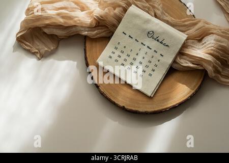 Draufsicht des Leinenstoffkalenders mit Oktobermonat, transparentem Stoff, Holzscheibe, heller Hintergrund, Kopierraum, ästhetische weiche Schatten Stockfoto