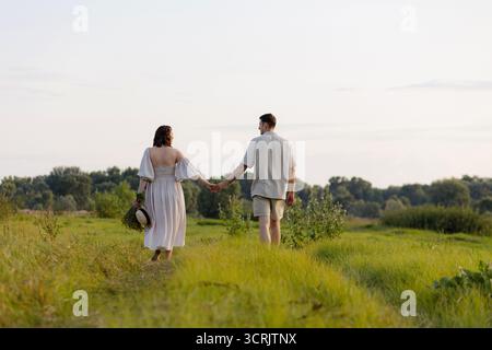 Ein Paar schlendert Hand in Hand durch eine üppige Wiese, umgeben von der Schönheit der Natur unter einem sanften Abendhimmel. Stockfoto