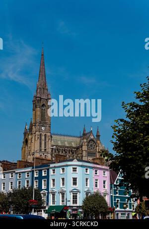 COBH, IRLAND - 15. August 2025: Die St. Colman's Cathedral erhebt sich über den farbenfrohen Geschäften und Pubs von Cobh und zieht Touristen an das bezaubernde Wasserfro der Stadt Stockfoto