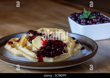 Nahaufnahme goldener Waffeln mit frischen Hucklebeeren, Beerenmarmelade und einem Klecks Butter auf einem rustikalen Teller. Stockfoto