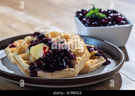 Nahaufnahme goldener Waffeln mit frischen Hucklebeeren, Beerenmarmelade und einem Klecks Butter auf einem rustikalen Teller. Stockfoto