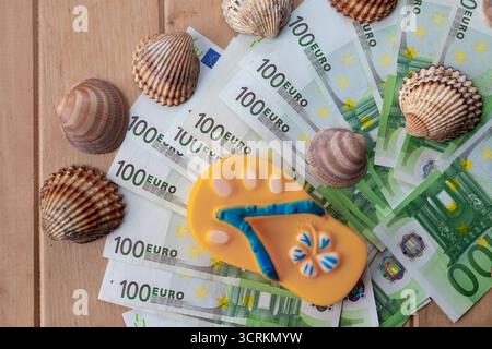 Stapel mit 100-Euro-Banknoten, die mit Flip-Flop und Muscheln auf Holztisch dekoriert sind. Konzept des Sommerurlaubsbudgets, Badeurlaubskosten, tra Stockfoto