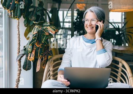 Fröhliche, reife Frau, die am Telefon spricht, während sie an einem Laptop arbeitet, in einem gemütlichen Café mit Pflanzen Stockfoto