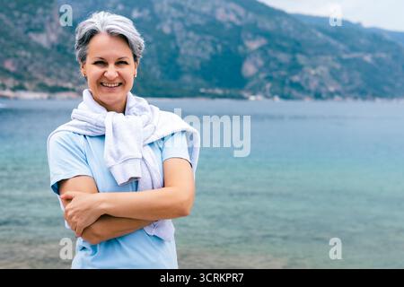 Glückliche Senior Woman by the Sea in hellblauem Hemd und Pullover, die Urlaub in der Natur genießen Stockfoto