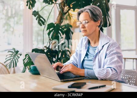 Konzentrierte, reife Frau, die an einem freiberuflichen Laptop-Projekt in einem gemütlichen, modernen Arbeitsbereich mit natürlichem Licht arbeitet Stockfoto