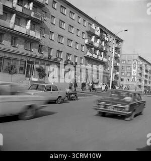 Ein dynamisches Schwarz-weiß-Archivfoto aus dem Jahr 1976, das eine geschäftige Straßenszene in der Chubarya-Straße in Sloviansk, ukrainische SSR, aufzeichnet. Das Bild ist voller klassischer sowjetischer Verkehrsmittel, wobei der berühmte VAZ-2101 „Zhiguli“, der als Stadttaxi arbeitet, und ein öffentlicher Bus PAZ-672 zu sehen sind. Das Geschehen entfaltet sich vor typischen fünfstöckigen Chruschtschjowka-Wohngebäuden, deren Fassaden mit festlichen Bannern zum 300. Jahrestag der Stadt geschmückt sind. Dies ist eine lebendige, atmosphärische Momentaufnahme der urbanen Mobilität und des Alltags in der UdSSR der 1970er Jahre. Stockfoto