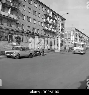 Ein dynamisches Schwarz-weiß-Archivfoto aus dem Jahr 1976, das eine geschäftige Straßenszene in der Chubarya-Straße in Sloviansk, ukrainische SSR, aufzeichnet. Das Bild ist voller klassischer sowjetischer Verkehrsmittel, wobei der berühmte VAZ-2101 „Zhiguli“, der als Stadttaxi arbeitet, und ein öffentlicher Bus PAZ-672 zu sehen sind. Das Geschehen entfaltet sich vor typischen fünfstöckigen Chruschtschjowka-Wohngebäuden, deren Fassaden mit festlichen Bannern zum 300. Jahrestag der Stadt geschmückt sind. Dies ist eine lebendige, atmosphärische Momentaufnahme der urbanen Mobilität und des Alltags in der UdSSR der 1970er Jahre. Stockfoto