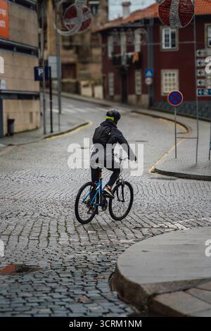 Rückansicht eines Radfahrers in schwarzer Jacke und Helm, der auf einer steilen Kopfsteinpflasterstraße in der Stadt Porto, Nordportugal, bergauf fährt Stockfoto