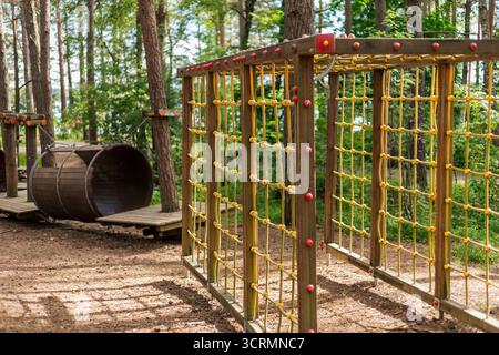 Ein Klettergerüst mit bunten Netzen steht in einem Waldpark neben einem Holzfass zum Spielen für Kinder. Üppiges Grün und Bäume schaffen es Stockfoto