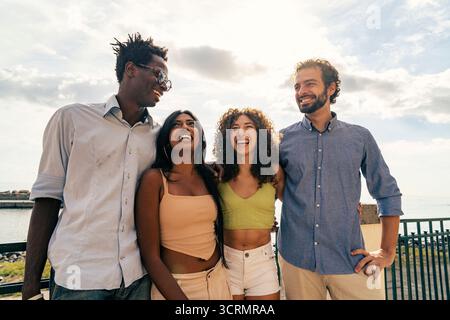 Eine fröhliche multikulturelle Gruppe von vier jungen Freunden steht zusammen und lacht und genießt einen schönen Sommerurlaub an der Küste. Stockfoto