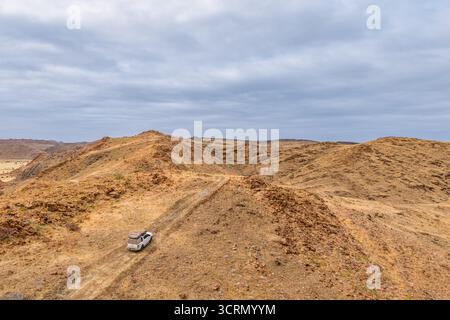 Aus der Vogelperspektive mit Blick auf ein einsames weißes Fahrzeug, das auf einem schroffen Schotterweg durch felsige, trockene Hügel navigiert, Twyfelfontein, Damaraland, Namibia. Stockfoto