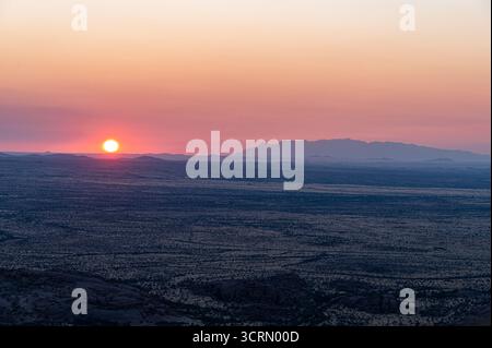 Leuchtende orange Sonne untergeht über einer weiten, flachen, trockenen Ebene unter einem rosa und violetten Abendhimmel mit fernen Bergen am Horizont, Damaraland, Namibia. Stockfoto