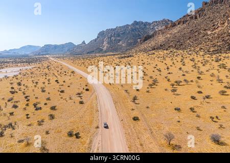 Standardmäßig aus der Luft ein einsames Fahrzeug, das gerade unbefestigte Straßen durch die goldenen Erongo-Ebenen fährt, und ein dramatisches felsiges Bergmassiv, das steil aus dem trockenen sa steigt Stockfoto
