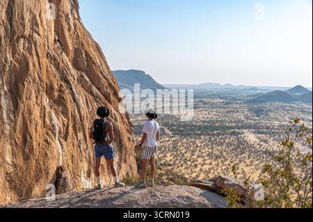 Zwei Wanderer stehen auf felsigen Klippen bei Erongo, Rucksäcke ruhen auf Granitfelsen, während sie über die weite namibische Wüstenebene, Namibia, blicken Stockfoto