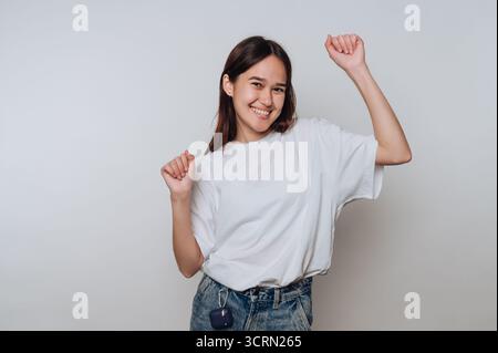Junge Frau mit braunen Haaren tanzt bei Tageslicht fröhlich in lässiger Kleidung vor einem schlichten Hintergrund Stockfoto