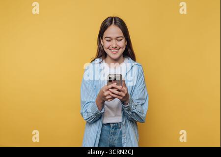 Eine lächelnde junge Frau mit einem gestreiften Hemd interagiert mit ihrem Smartphone vor einem hellgelben Hintergrund Stockfoto