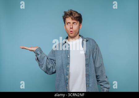 Junger Mann mit lässigem Outfit zeigt überraschenden Ausdruck auf blauem Hintergrund, während er mit der Hand Gesten zeigt Stockfoto