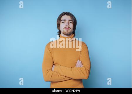 Ein selbstbewusster junger Mann in gelbem Rollkragenpullover steht vor blauem Hintergrund, während die Arme beim Fotoshooting in der Halle überkreuzt werden Stockfoto