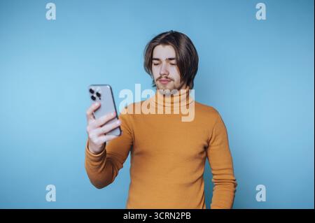 Ein junger Mann, der einen Rollkragenpullover trägt, sieht sein Smartphone vor einem blauen Hintergrund an Stockfoto