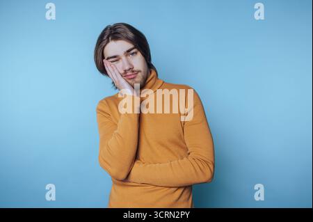Ein junger Mann in einem gelben Pullover posiert nachdenklich vor blauem Hintergrund und hält einen Moment der Introspektion und Besinnung fest Stockfoto