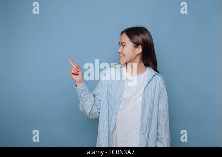 Eine junge Frau steht vor einem hellblauen Hintergrund, trägt ein gestreiftes Hemd und ein schlichtes weißes T-Shirt. Sie lächelt und zeigt nach rechts und überträgt die Zehnte Stockfoto