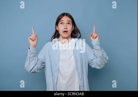 Eine junge Frau, die ein helles gestreiftes Hemd und ein weißes Oberteil trägt, zeigt Überraschung und Neugier, während sie mit beiden Fingern nach oben auf einen blauen Rücken zeigt Stockfoto