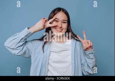 Eine junge Frau steht vor einem sanften blauen Hintergrund, blitzt ein Lächeln und macht mit beiden Händen Friedenszeichen, die eine fröhliche und entspannte Haltung zeigen Stockfoto