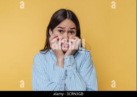 Eine junge Frau erscheint sichtbar ängstlich, ihre Hände greifen ihr Gesicht und einen besorgten Ausdruck. Der hellgelbe Hintergrund erhöht die Intensität von Stockfoto