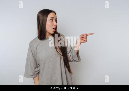 Eine junge Frau mit langen Haaren, die ein graues übergroßes Hemd trägt, zeigt Überraschung und zeigt zur Seite. Sie wirkt engagiert und ausdrucksstark in einer minimalistischen Stockfoto
