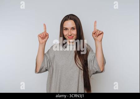 Eine junge Frau steht in einer hellen Umgebung, lächelt weit und hebt ihre Hände mit den Fingern nach oben. Ihre lässige Kleidung und ihre fröhliche Ausstrahlung Stockfoto