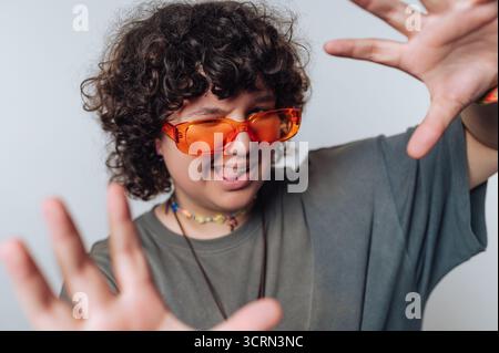 Eine junge Person mit lockigem Haar zeigt eine ausgelassene Persönlichkeit durch verspielte Handgesten, in leuchtend orange Sonnenbrille und einem lässigen Shi Stockfoto