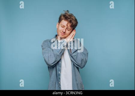 Ein junger Mann mit lockigen Haaren lächelt, während er eine schlafende Pose nachahmt und eine fröhliche Stimmung zeigt. Der sanfte blaue Hintergrund verstärkt die verspielte Atmosphäre, Stockfoto