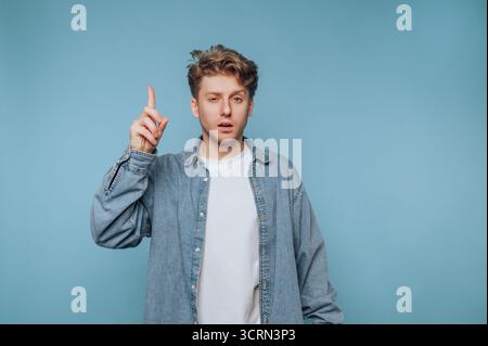 Ein junger Mann steht mit erhobenem Finger vor einem hellblauen Hintergrund und erscheint tief in Gedanken. Er trägt lässig eine Jeansjacke und weiß Stockfoto