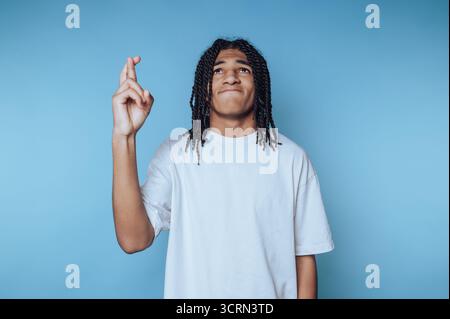 Ein junger Mensch steht mit hoffnungsvollem Ausdruck, mit gekreuzten Daumen, vor einem blauen Hintergrund. Die entspannte Haltung deutet auf Vorfreude oder Stockfoto