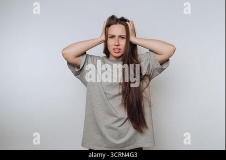 Eine junge Frau mit langen Haaren sieht gestresst aus und hält ihren Kopf mit beiden Händen vor einem schlichten Hintergrund. Stockfoto
