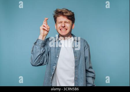 Ein junger Mann mit lockigen Haaren kreuzt mit geschlossenen Augen die Finger und drückt Hoffnung oder Vorfreude vor blauem Hintergrund aus. Stockfoto