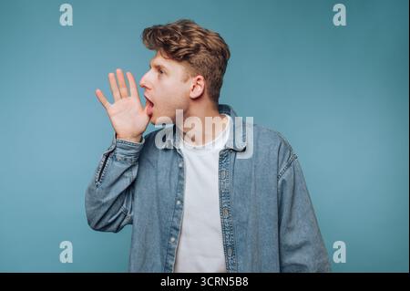 Junger Mann im Jeanshemd, der mit der Hand in Mundnähe vor blauem Hintergrund brüllt Stockfoto