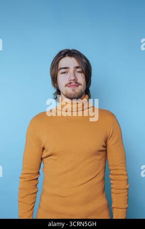 Junger Mann mit langen Haaren und Bart, der einen Senfkragen vor blauem Hintergrund trägt. Stockfoto
