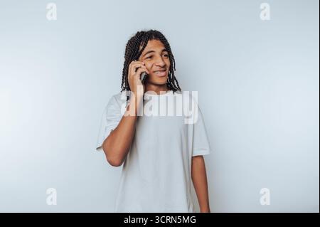 Junger Mann mit Dreadlocks, der auf einem Smartphone spricht, ein weißes T-Shirt trägt und vor einem weißen Hintergrund steht. Stockfoto