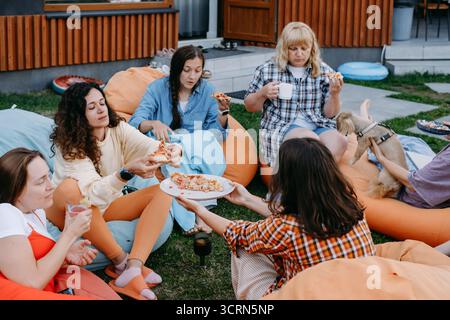 Freunde, die sich auf bunten Sitzsäcken im Freien entspannen und Essen und Getränke in zwangloser geselliger Atmosphäre mit einem kleinen Hund teilen. Frau informell Stockfoto