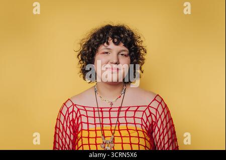 Eine Person mit lockigem Haar, die ein rotes Netztop über einem gelben Hemd trägt, lächelt vor gelbem Hintergrund. Stockfoto