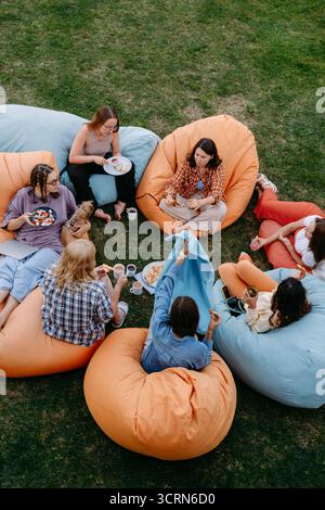 Freunde, die sich auf bunten Sitzsäcken im Freien entspannen und Essen und Getränke in zwangloser geselliger Atmosphäre mit einem kleinen Hund teilen. Frau informell Stockfoto