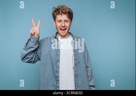 Porträt eines jungen Mannes zeigt ein Friedenszeichen während des Lächelns und strahlt positive Energie auf hellblauem Hintergrund aus Stockfoto